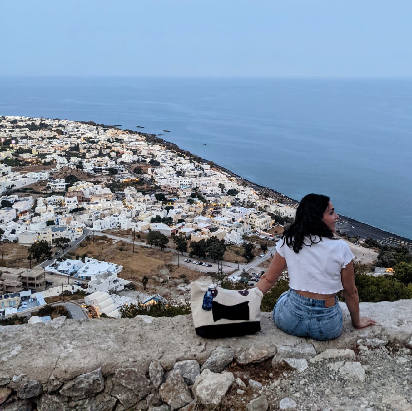 Alexa sits on the edge of a wall overlooking the village in Ancient Thira, Santorini, Greece. Her back is to the camera