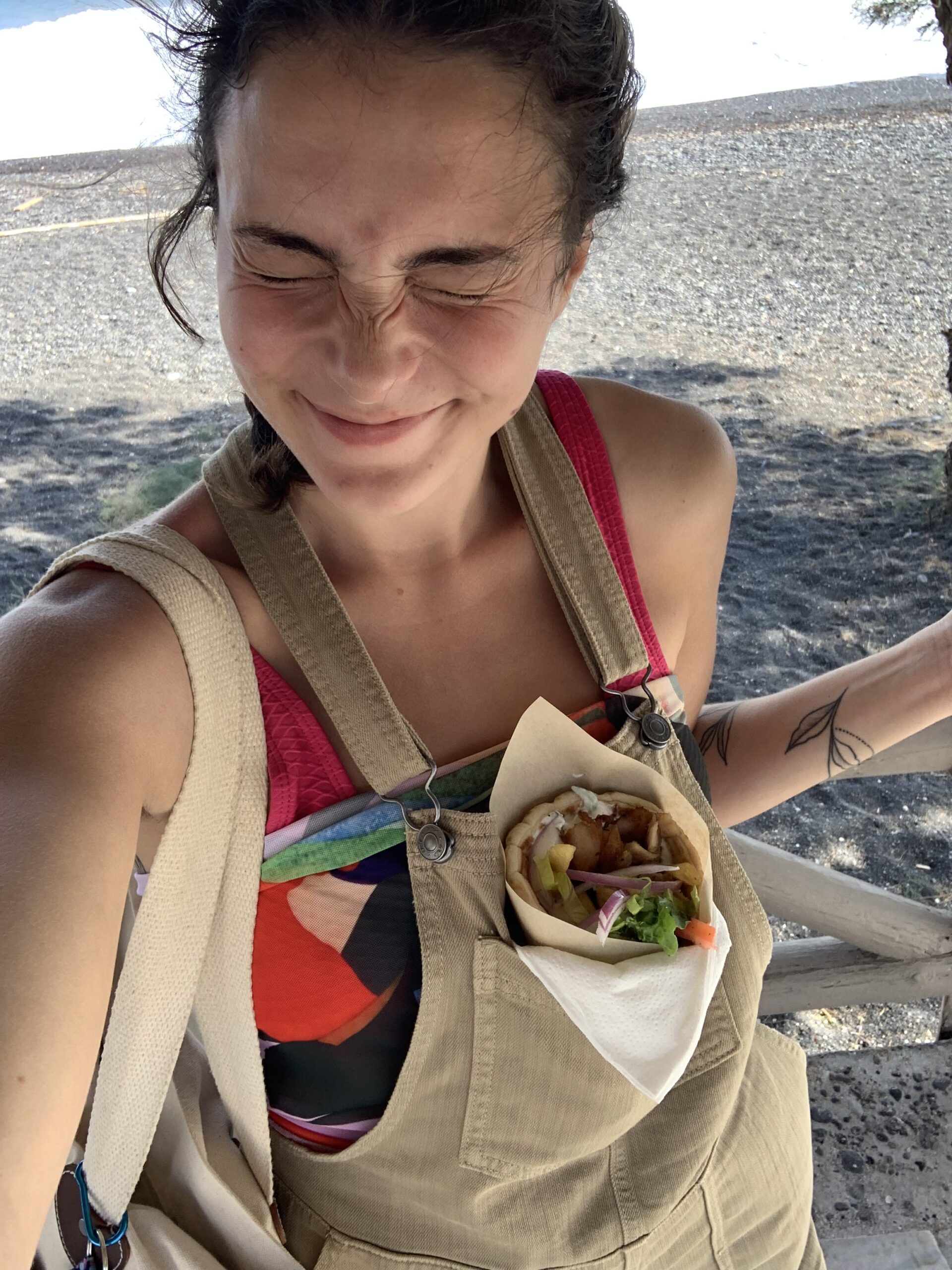 Alexa smiles with a gyro in the pocket of her overalls on the beach in Santorini, Greece