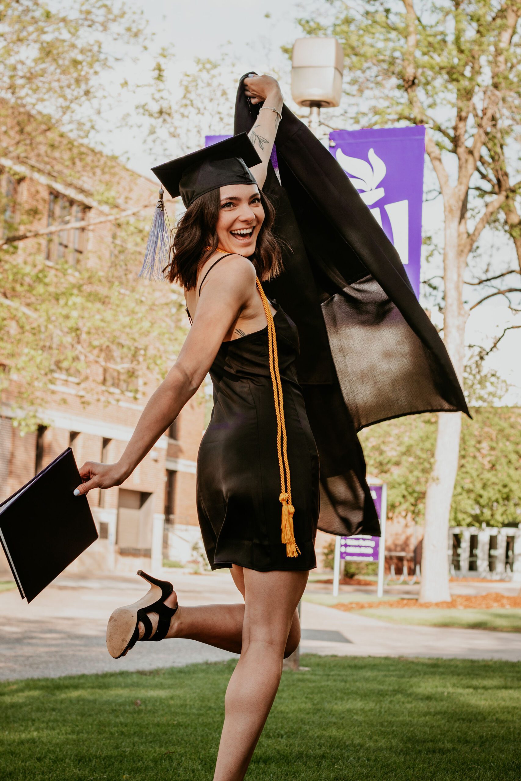 Alexa smiles and throws out her arms with her graduation gown in one hand and diploma in the other