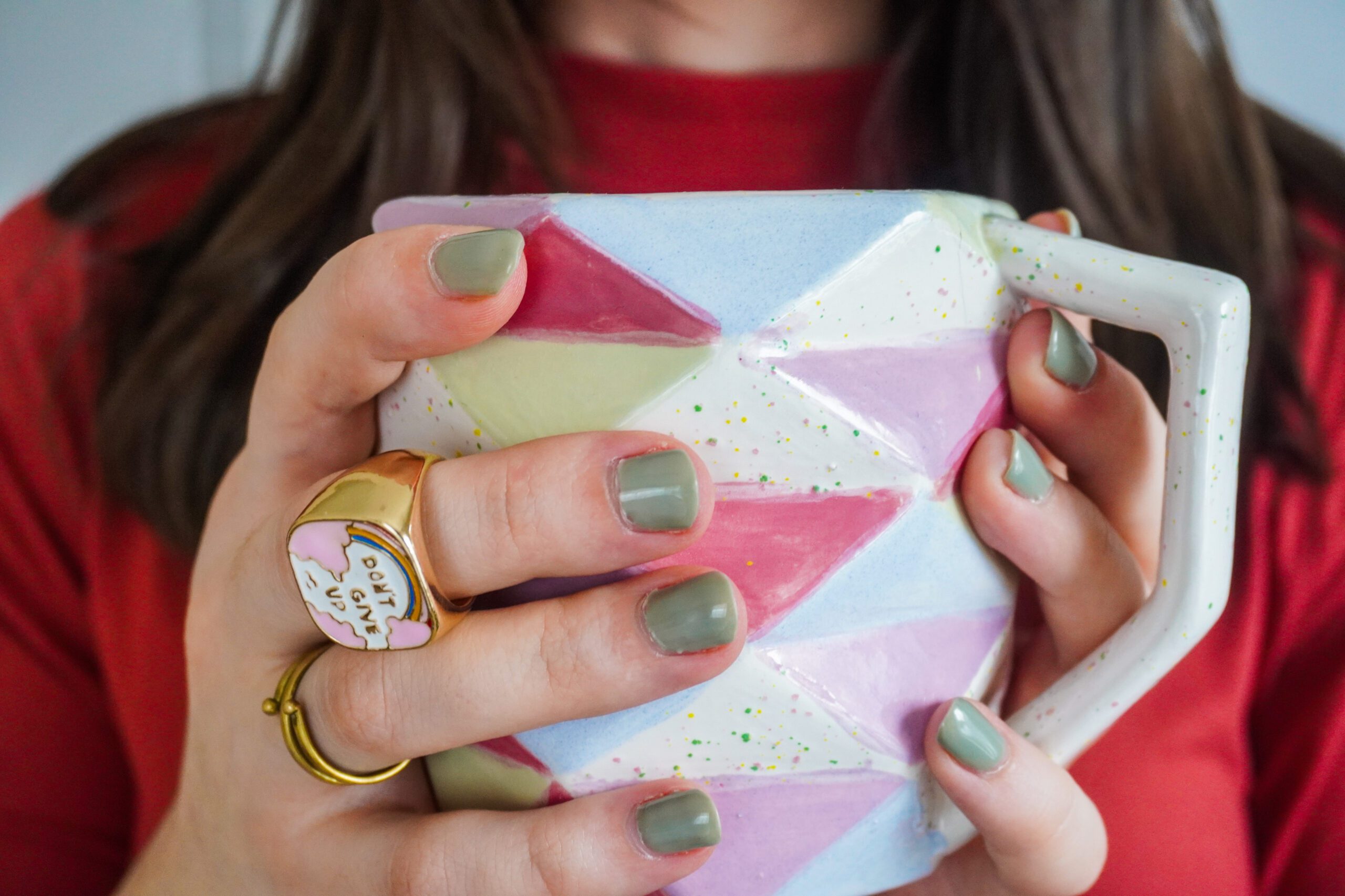 A geometric colorful coffee mug is held by Alexa. Her nails are painted green and she is wearing gold rings.