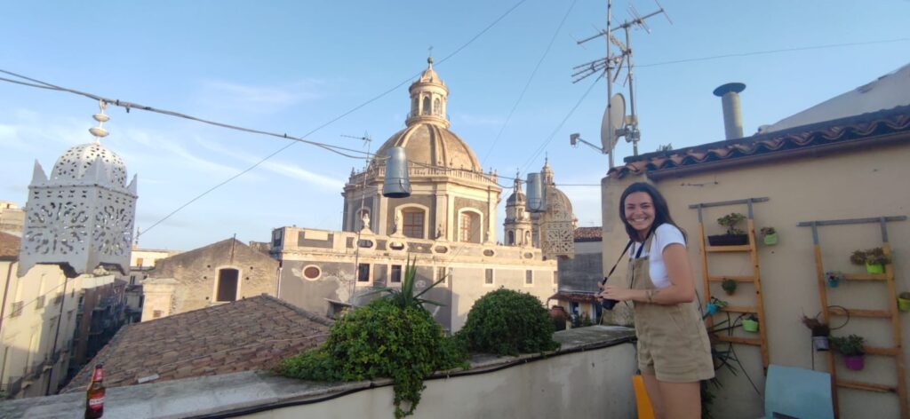 Alexa stands on a chair on a rooftop in Catania, Italy. She has a camera in her hands and is smiling.