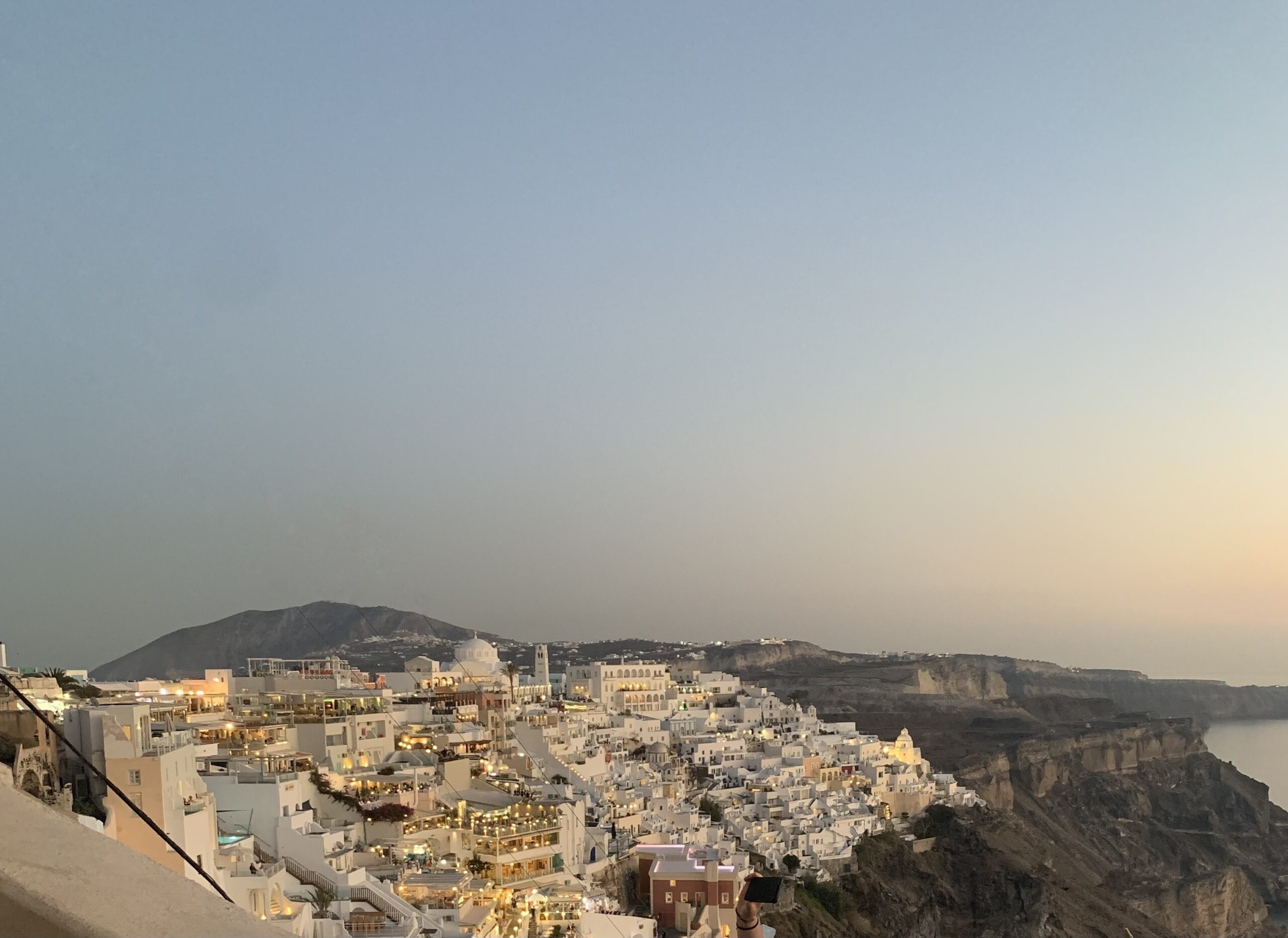 Warmly lit white houses cover a mountain side in Santorini at dusk.