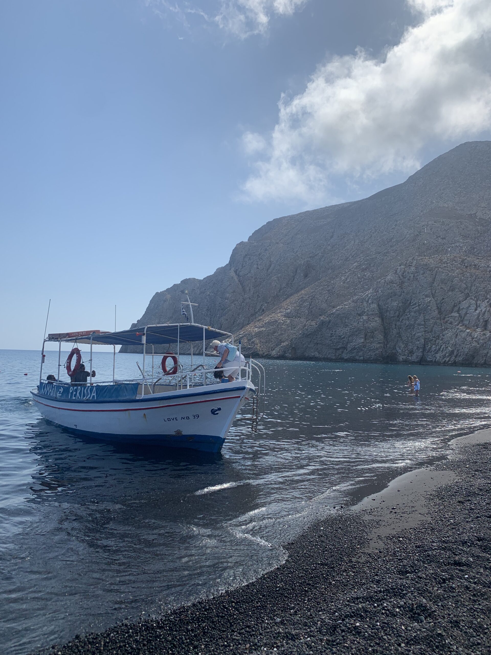 A small boat sits at the shore on the beach in Santorini, Greece. The boat has "LOVE NB 39" on its side.