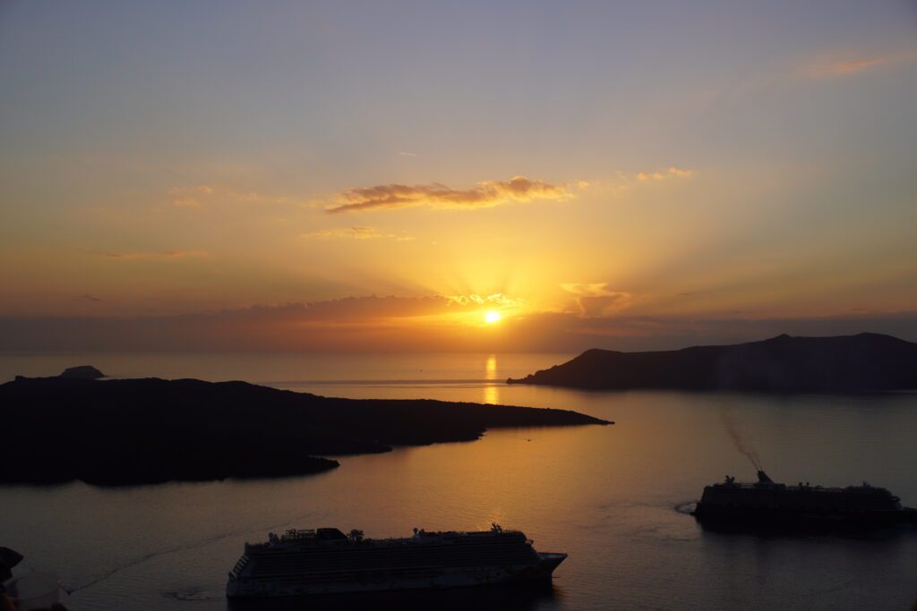 A sunset stretches over the ocean and silhouetted islands and cruise ships in Santorini, Greece.