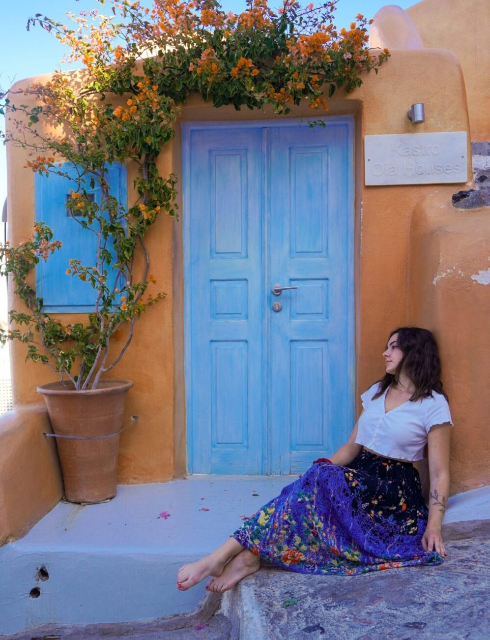 Alexa sits on the step in front of a blue door looking outward. She is wearing a white top and colorful skirt.