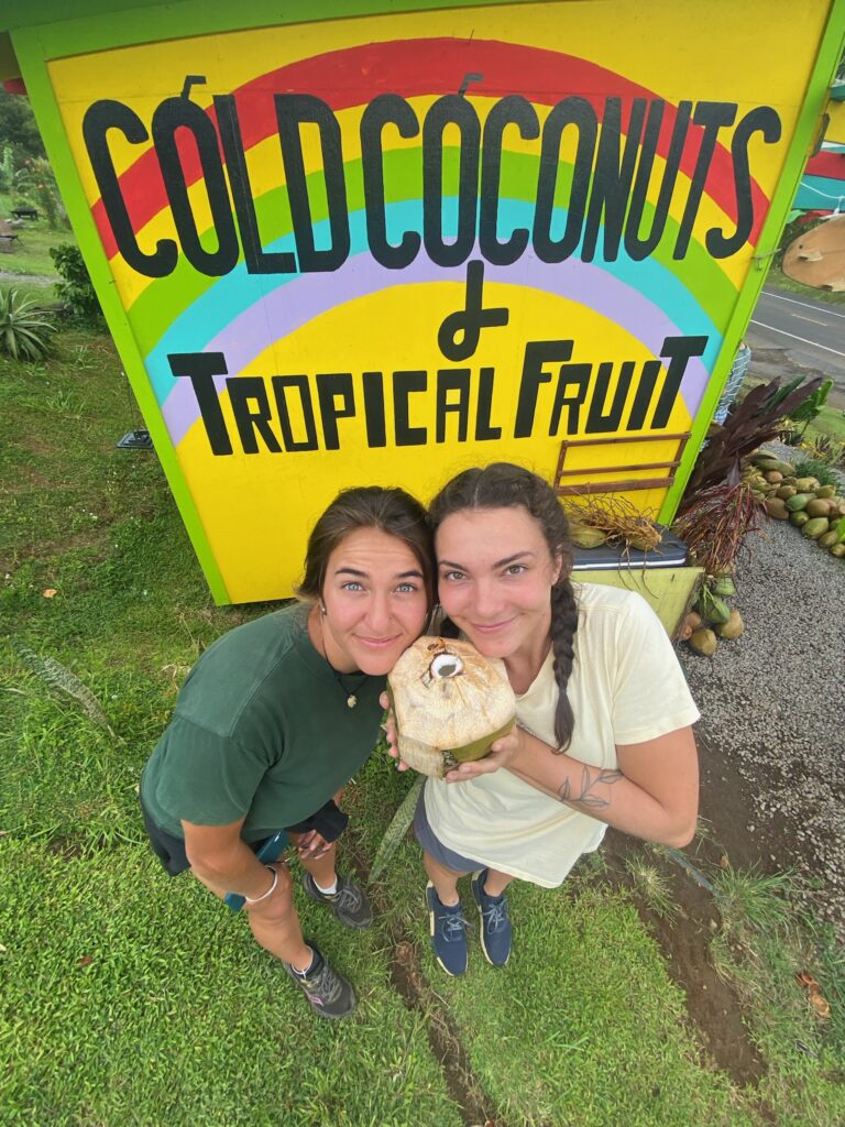 Two women standing in front of a sign that says cold coconuts tropical fruit.