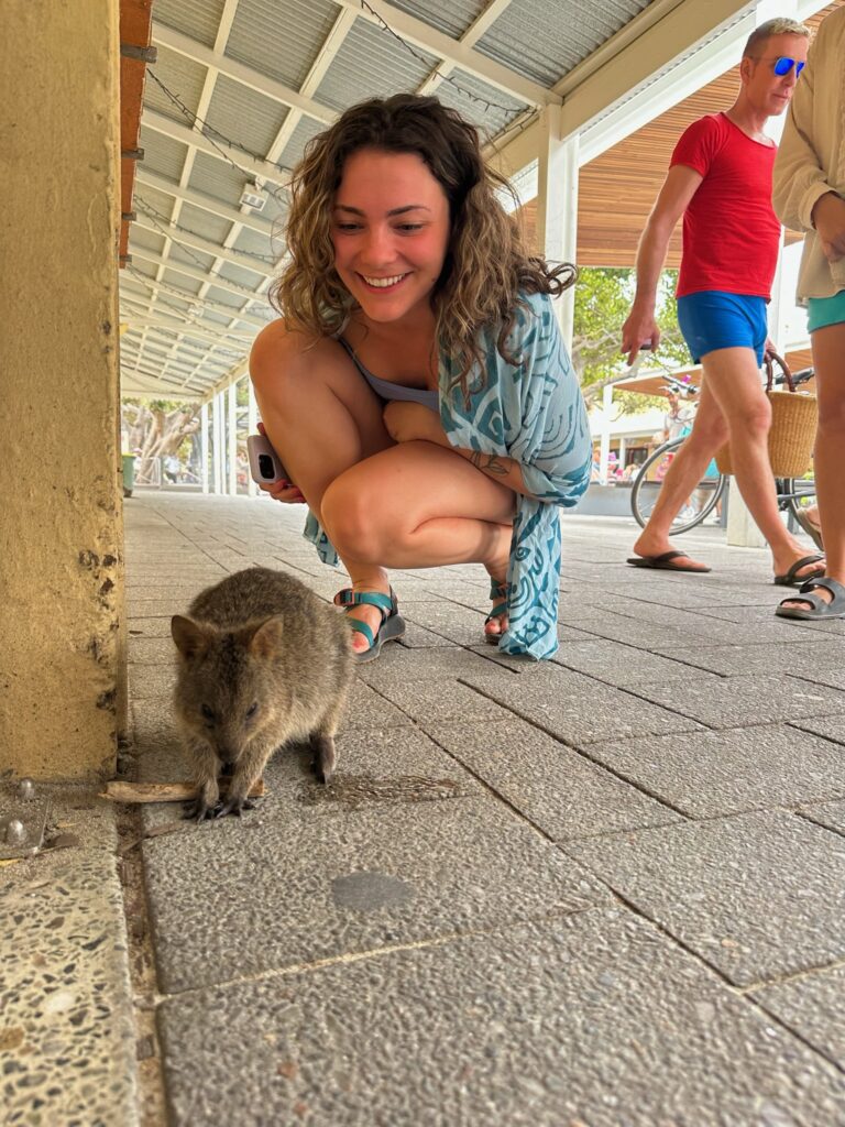 A woman crouching down smiling at a small quokka on a pedestrian walkway.