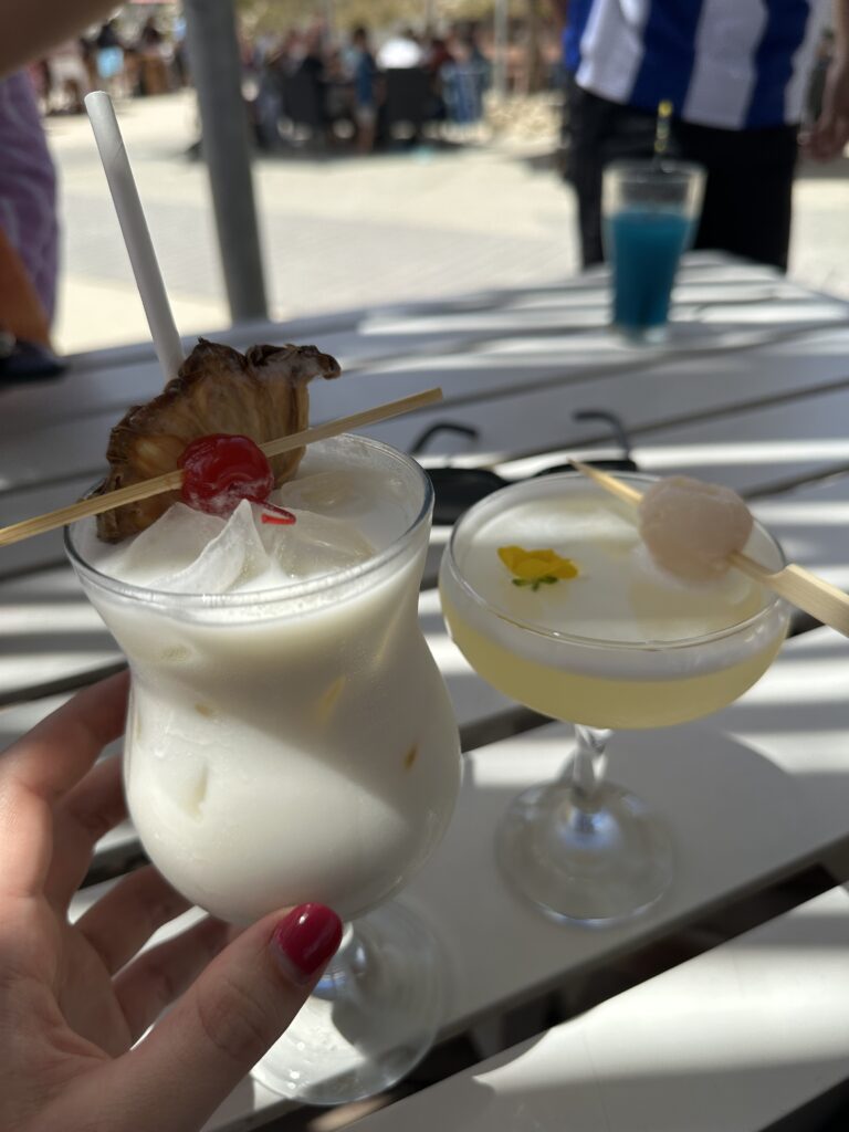 A person holding a creamy cocktail garnished with a cherry and a skewered piece of fruit, with another cocktail with a floral garnish in the background.