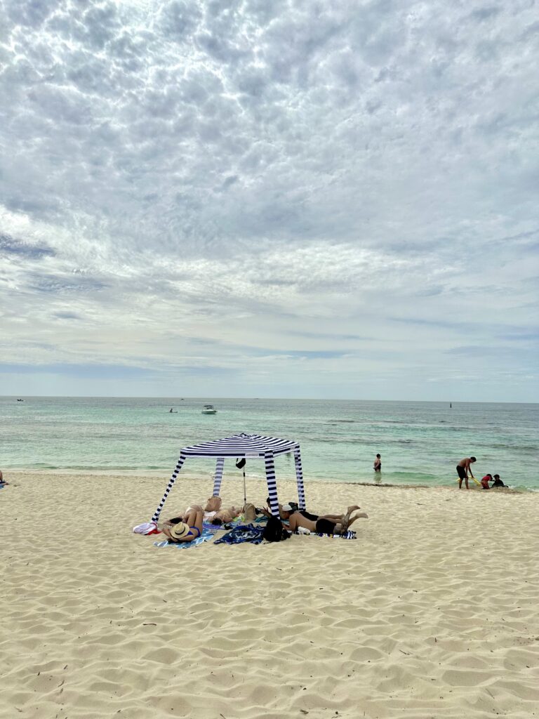 A shaded seating area with two people and a dog on a sandy beach, overlooking a calm sea and partly cloudy sky.