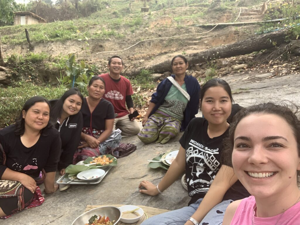 Group of seven people sitting on the ground outdoors, sharing a meal, with greenery and farmland in the background.