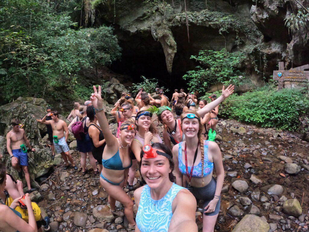 Group of cheerful people in swimwear posing for a selfie in front of a cave with lush green vegetation around at Khao Sok National Park.