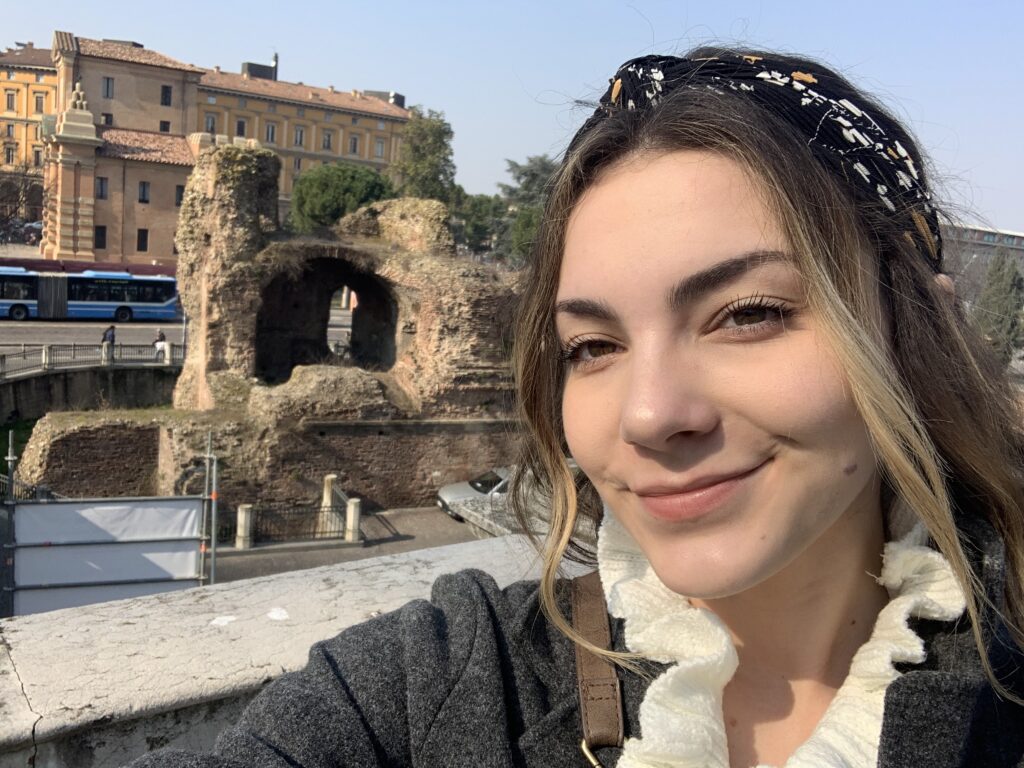 A young woman with a headband smiling at the camera, with ancient ruins and a blue bus in the background.