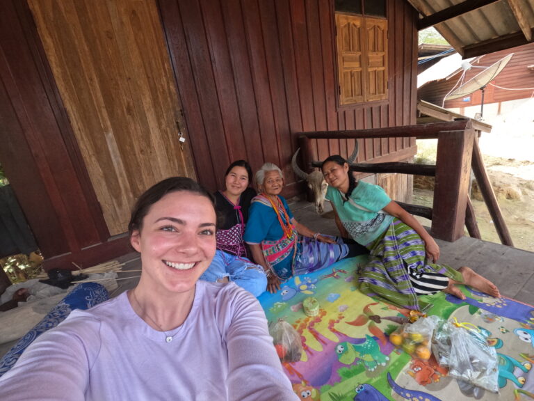 Four women of different ages smiling at the camera, sitting together on a mat outside a wooden house in a rural setting.