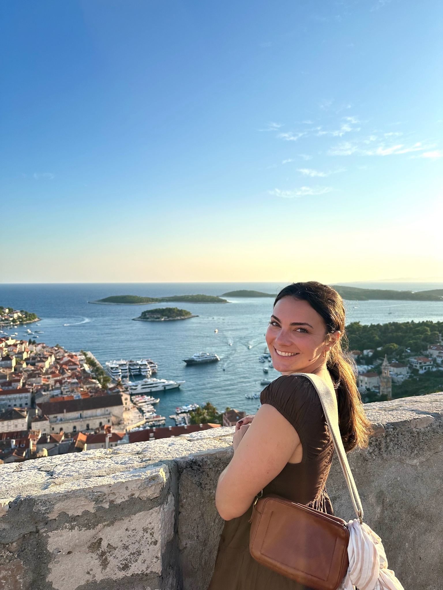 Person smiling while standing at a scenic overlook, with a coastal town and ocean in the background under a clear blue sky.