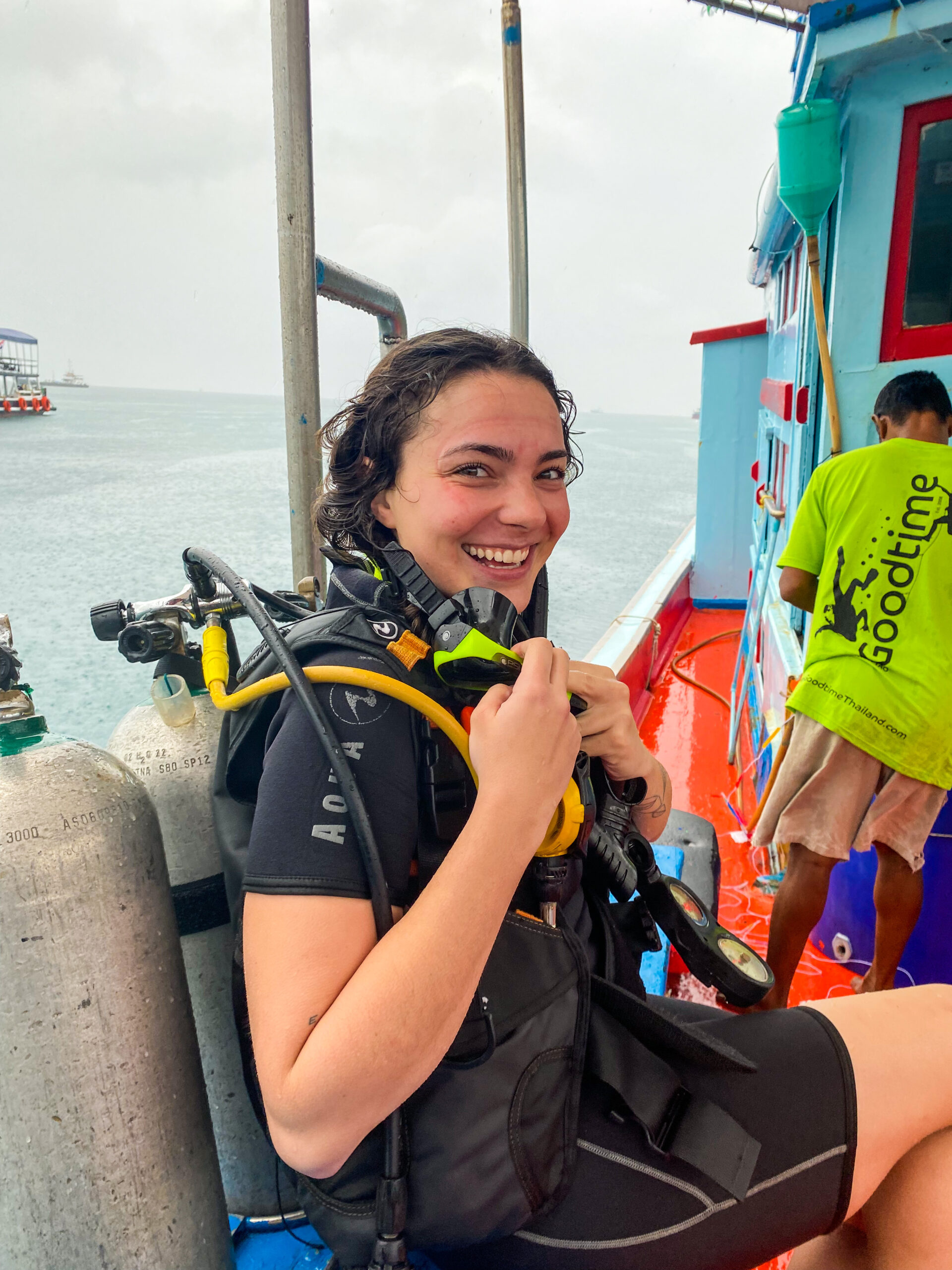 A person in diving gear sitting on a red and blue boat, smiling at the camera. Another person in a green shirt is adjusting equipment in the background.